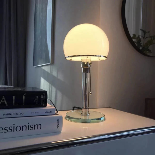 Bauhaus-inspired table lamp emitting warm light, placed on a white table with books and a mirror reflection.